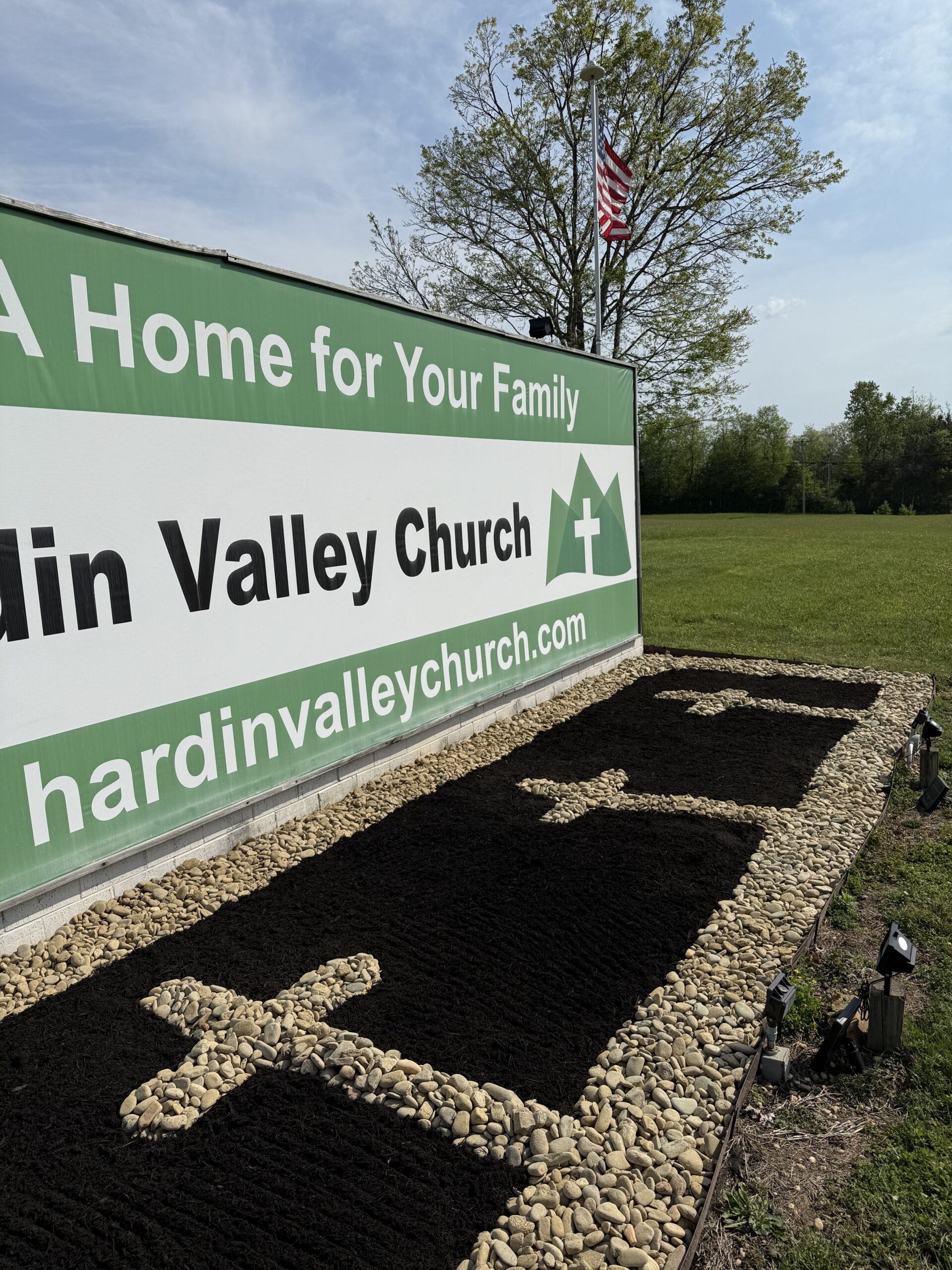 Sign for Hardin Valley Church next to a landscaped area with crosses made of rocks, bordered by pebbles and grass, with an American flag and trees in the background under a blue sky.