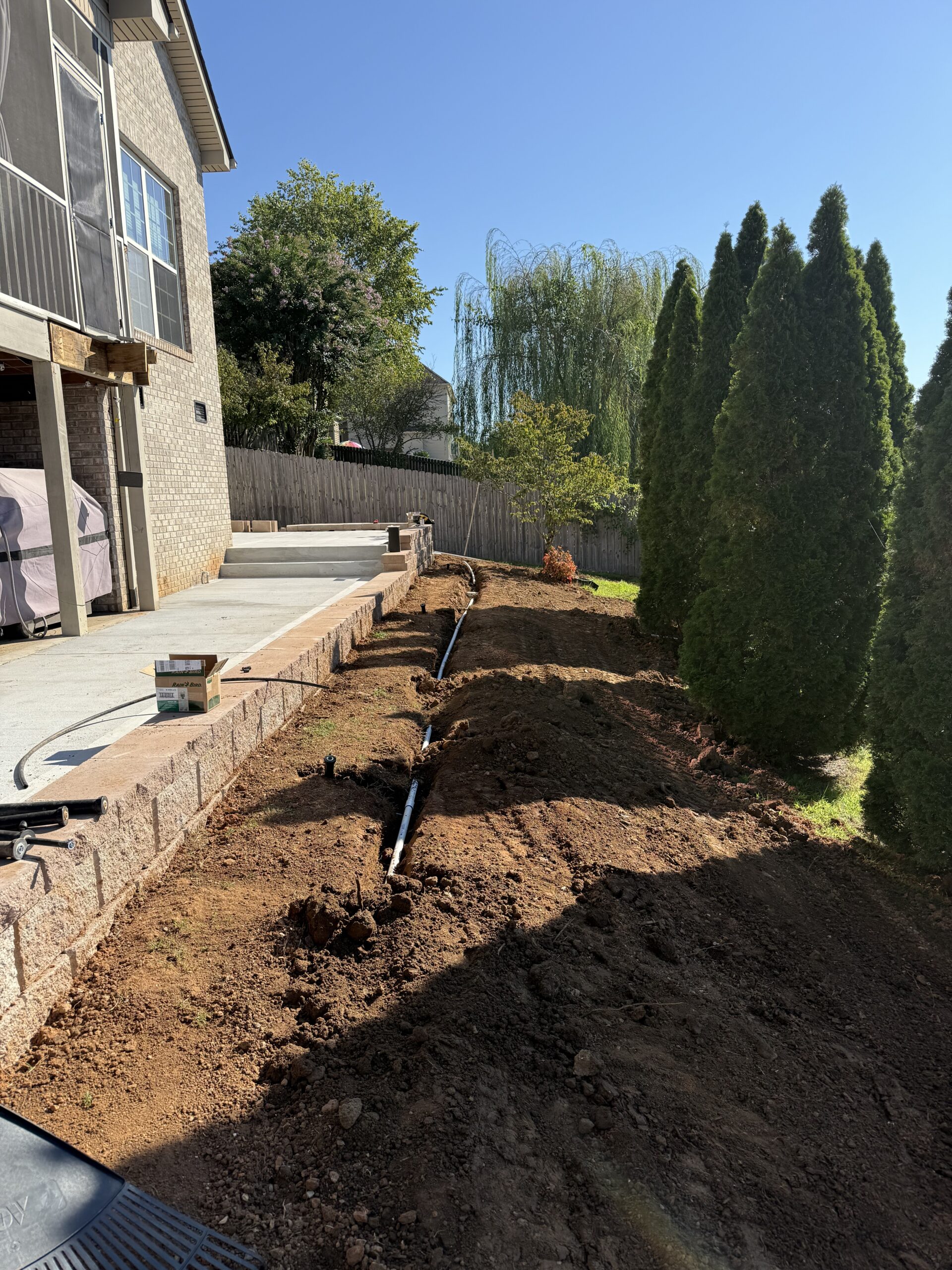 A backyard with freshly dug dirt, exposed pipes running along a retaining wall, and evergreen trees bordering the yard under a clear sky.