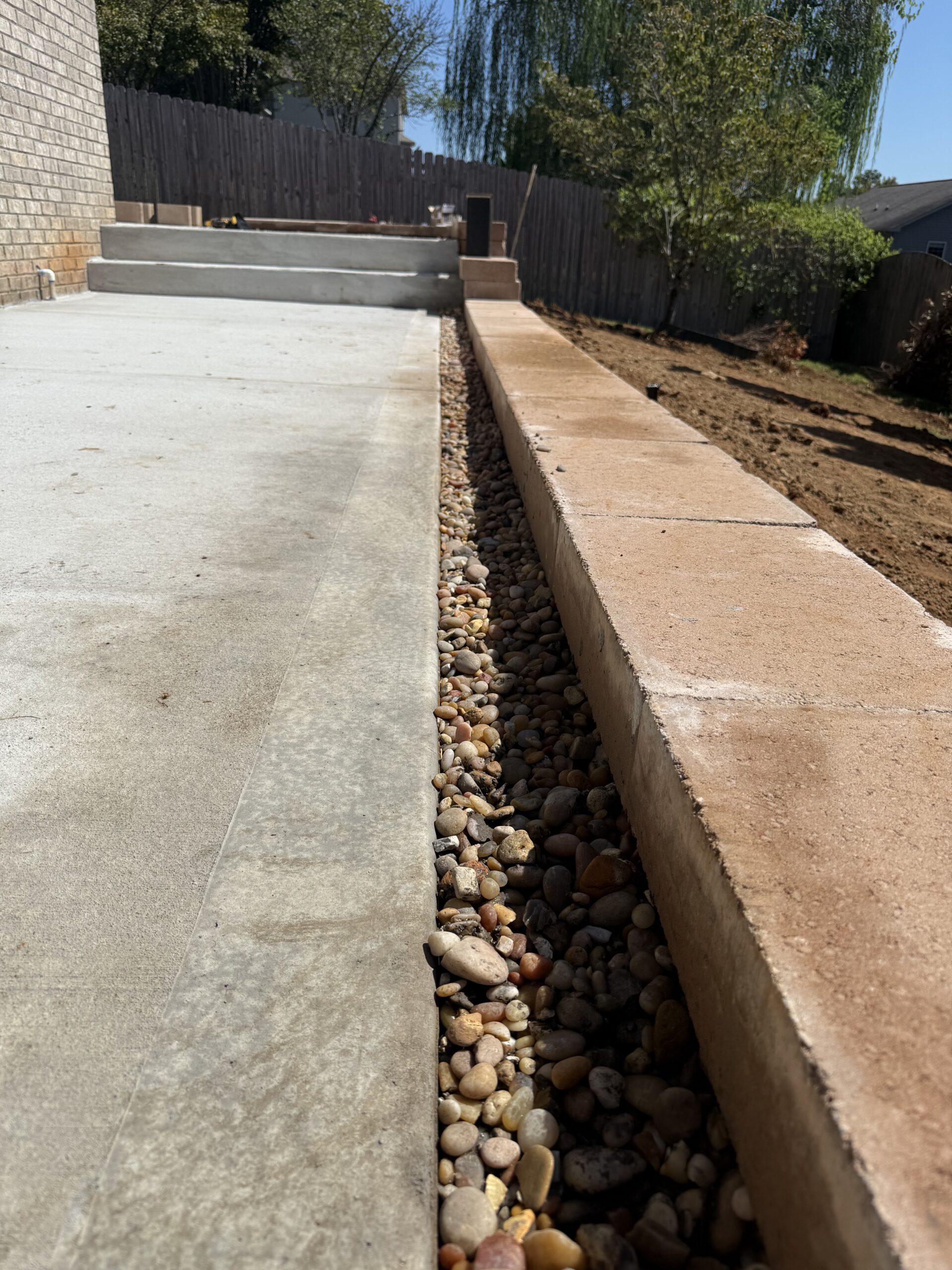 Close-up of a concrete patio next to a brick retaining wall with a narrow strip of decorative rocks in between, in a backyard under sunny skies.