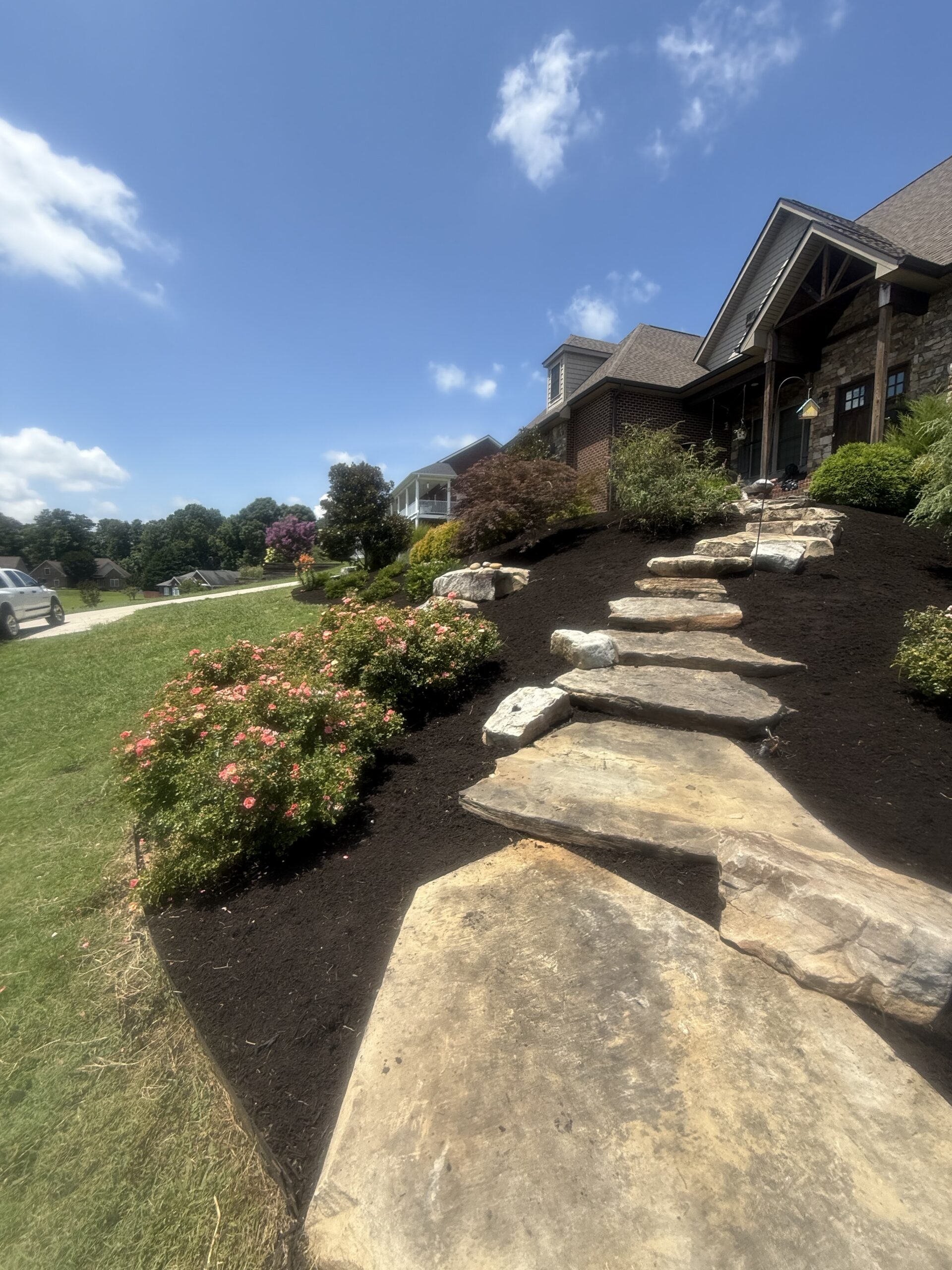 Stone steps lead up a landscaped incline with flowering bushes and mulch, next to a house under a blue sky with scattered clouds.
