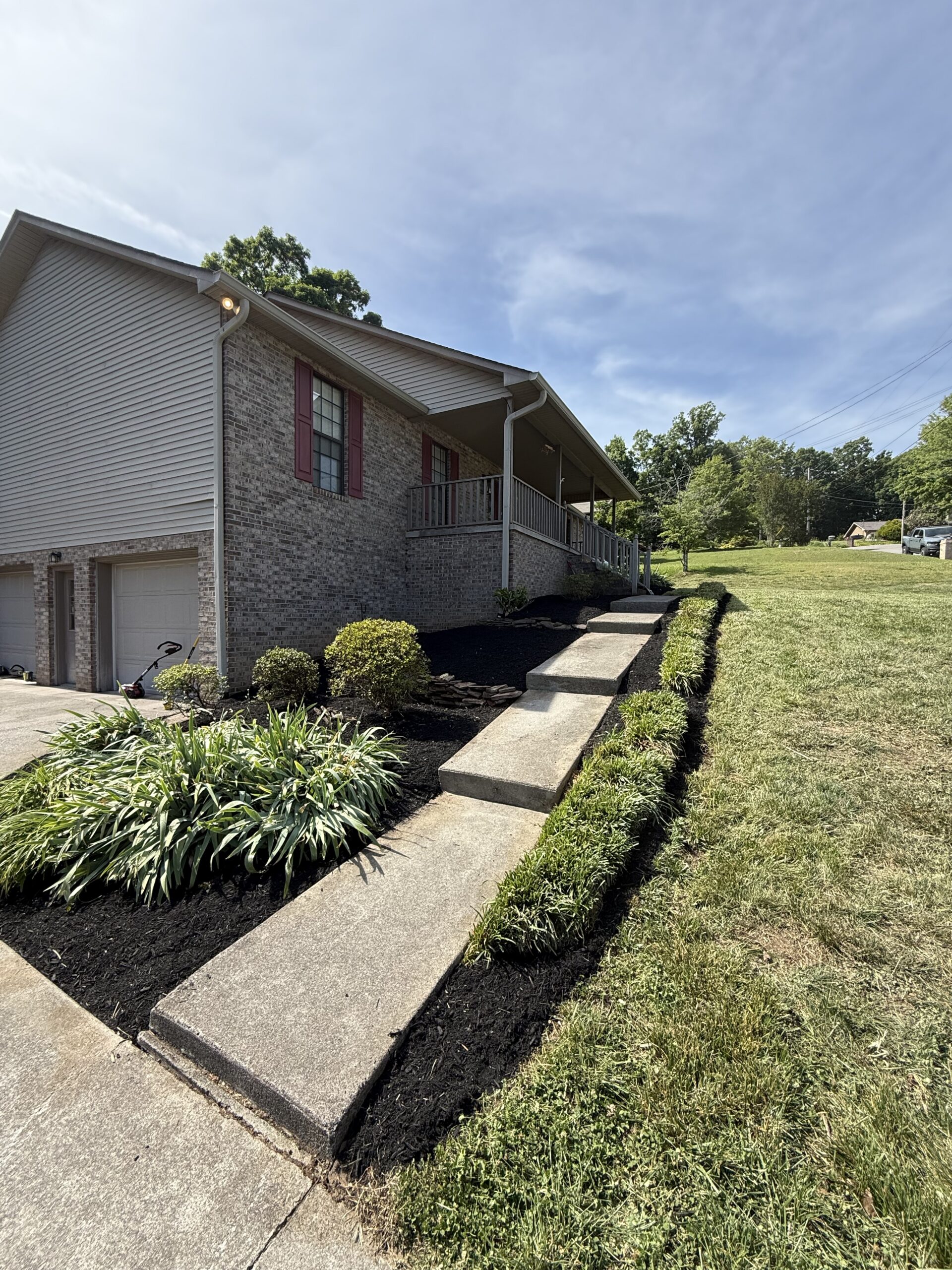 Concrete steps lead up to a covered porch of a brick house with red shutters, landscaped bushes, and a grassy yard on a sunny day.