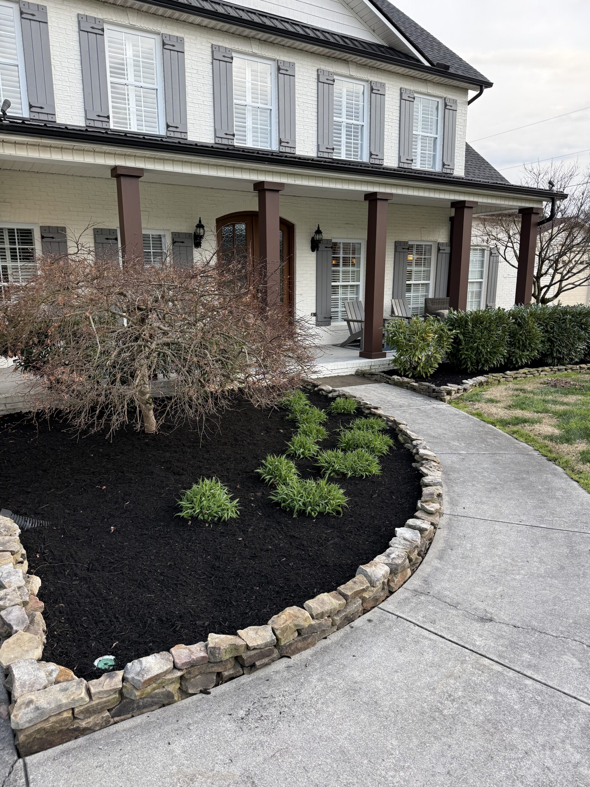 A stone-edged garden bed with black mulch and green plants sits beside a curved concrete walkway leading to a white brick house with brown columns and shutters.