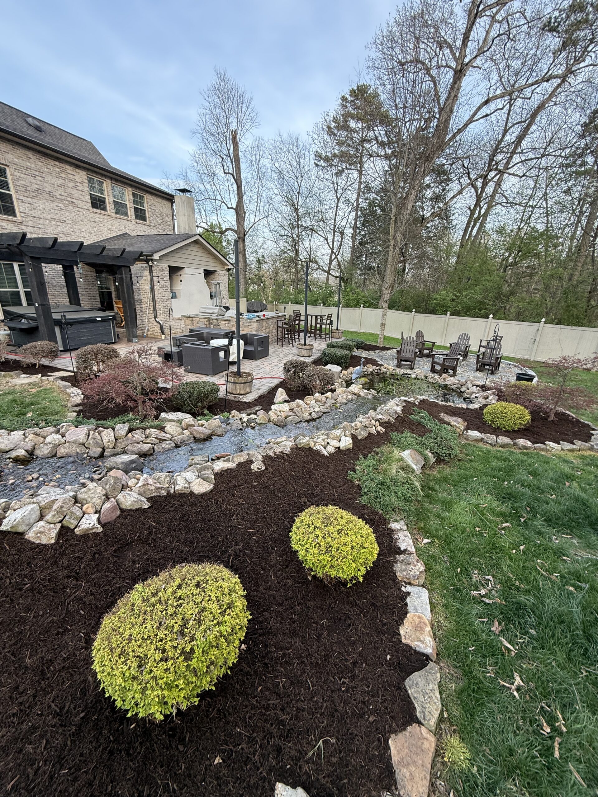 Backyard with landscaped garden, mulched flower beds, round shrubs, stone borders, a patio with outdoor seating, fire pit area, and surrounding trees.