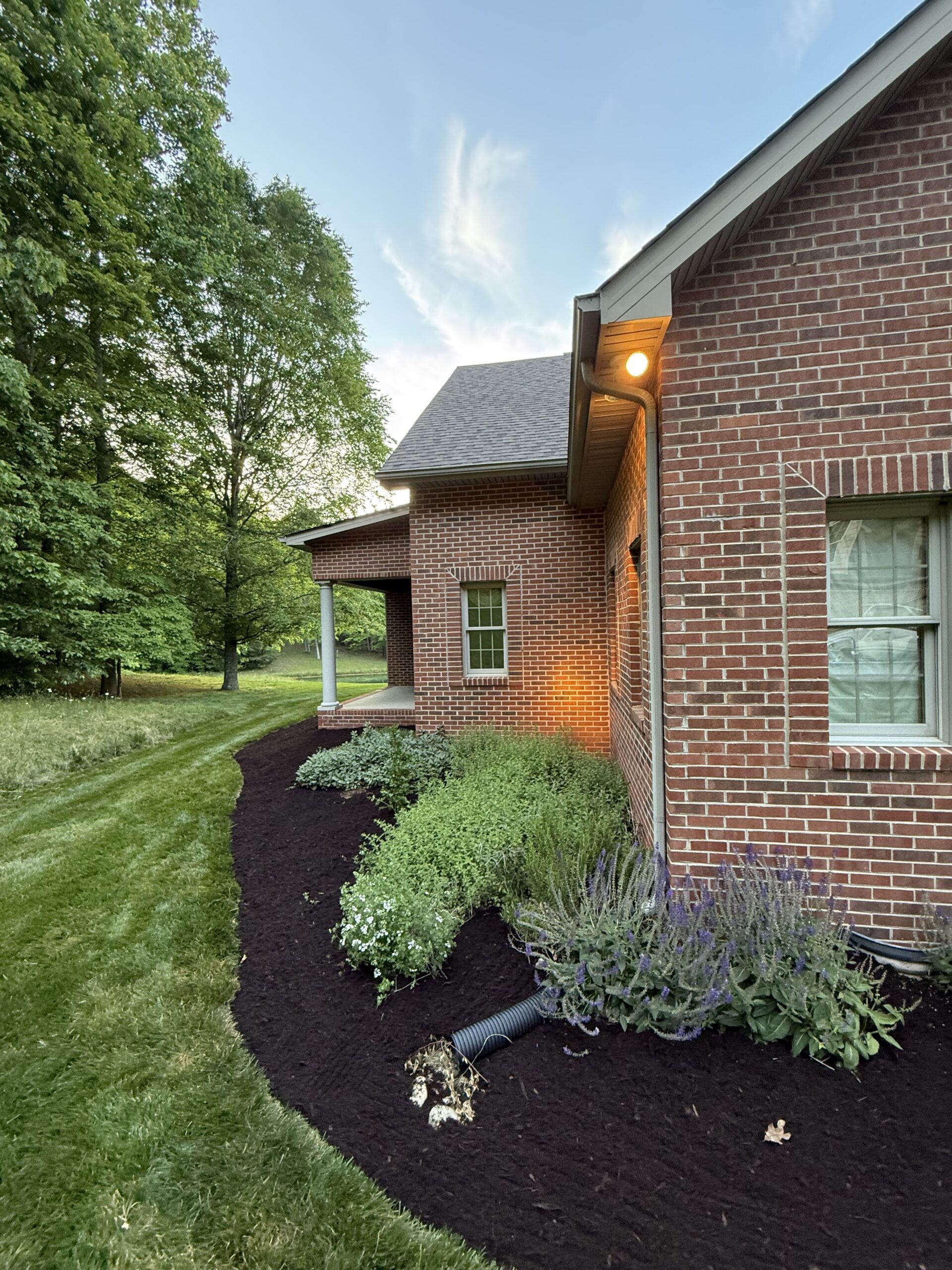 Red brick house with a covered porch, lit exterior light, and a landscaped garden bed with green plants and mulch beside a grassy lawn and trees.