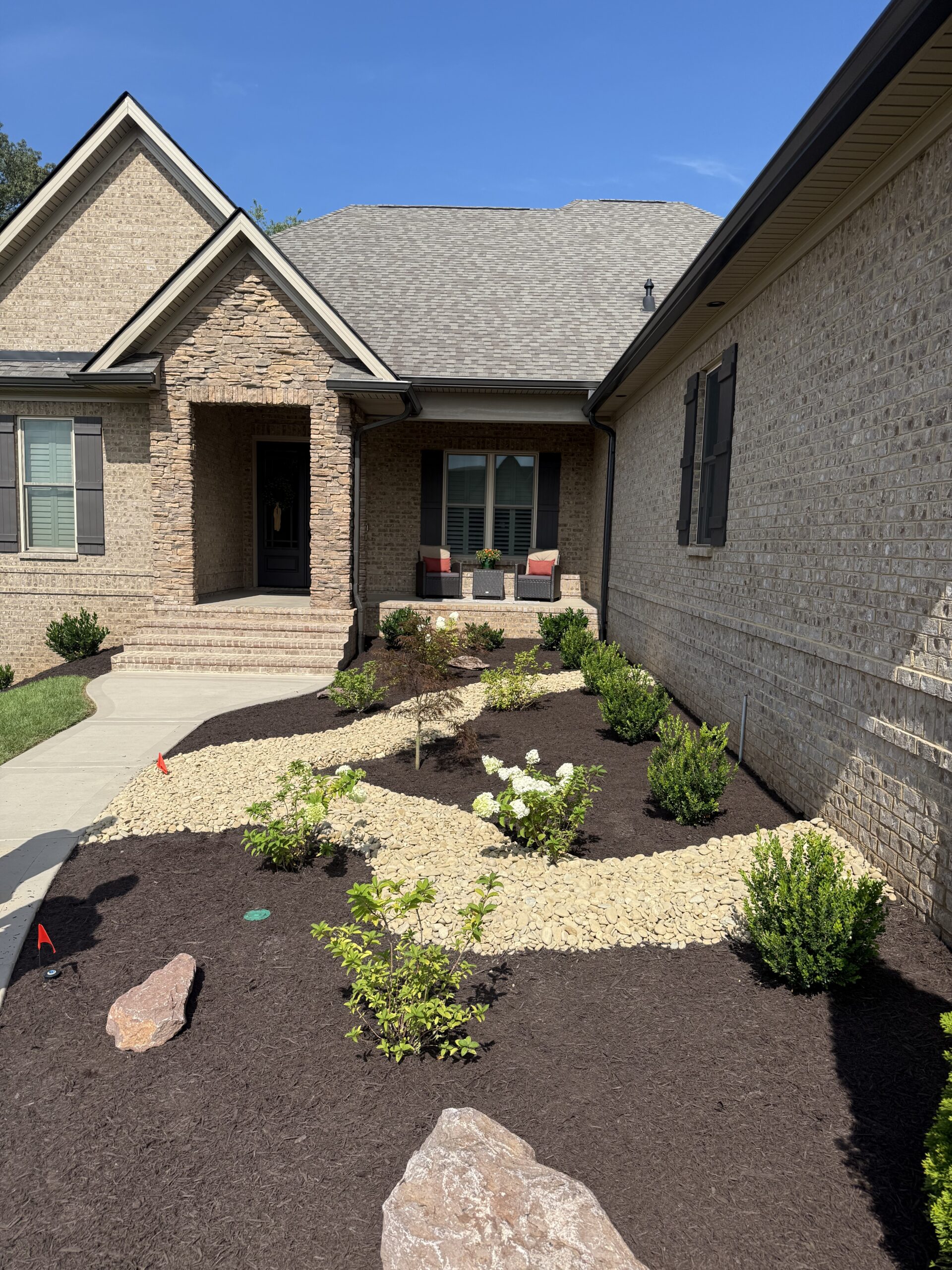 A landscaped front yard with mulch, decorative rocks, small shrubs, and a stone path leading to a brick house with a covered porch.