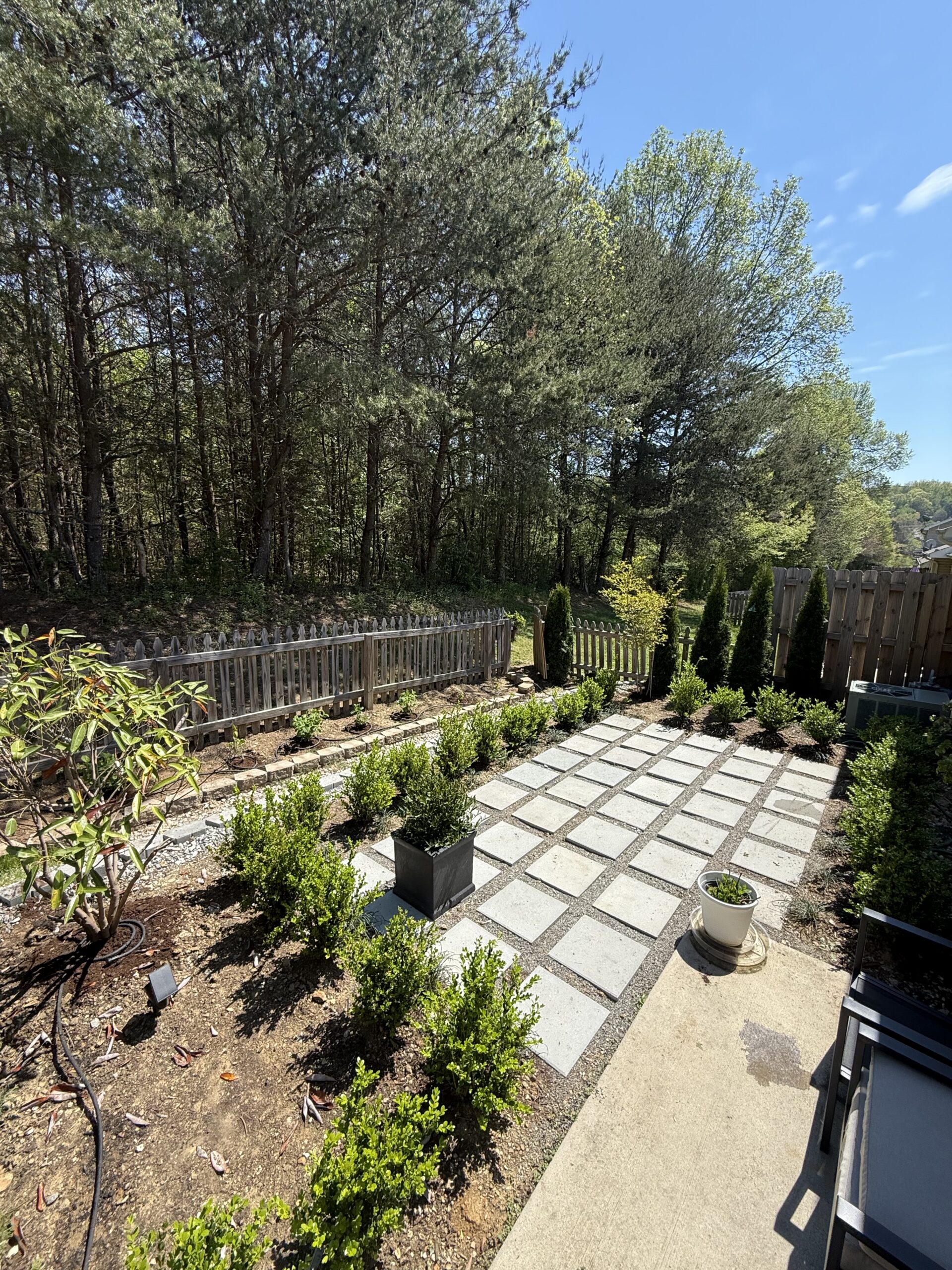 A backyard patio with square pavers, planter boxes, small shrubs, and a wooden fence, bordered by tall trees under a clear sky.